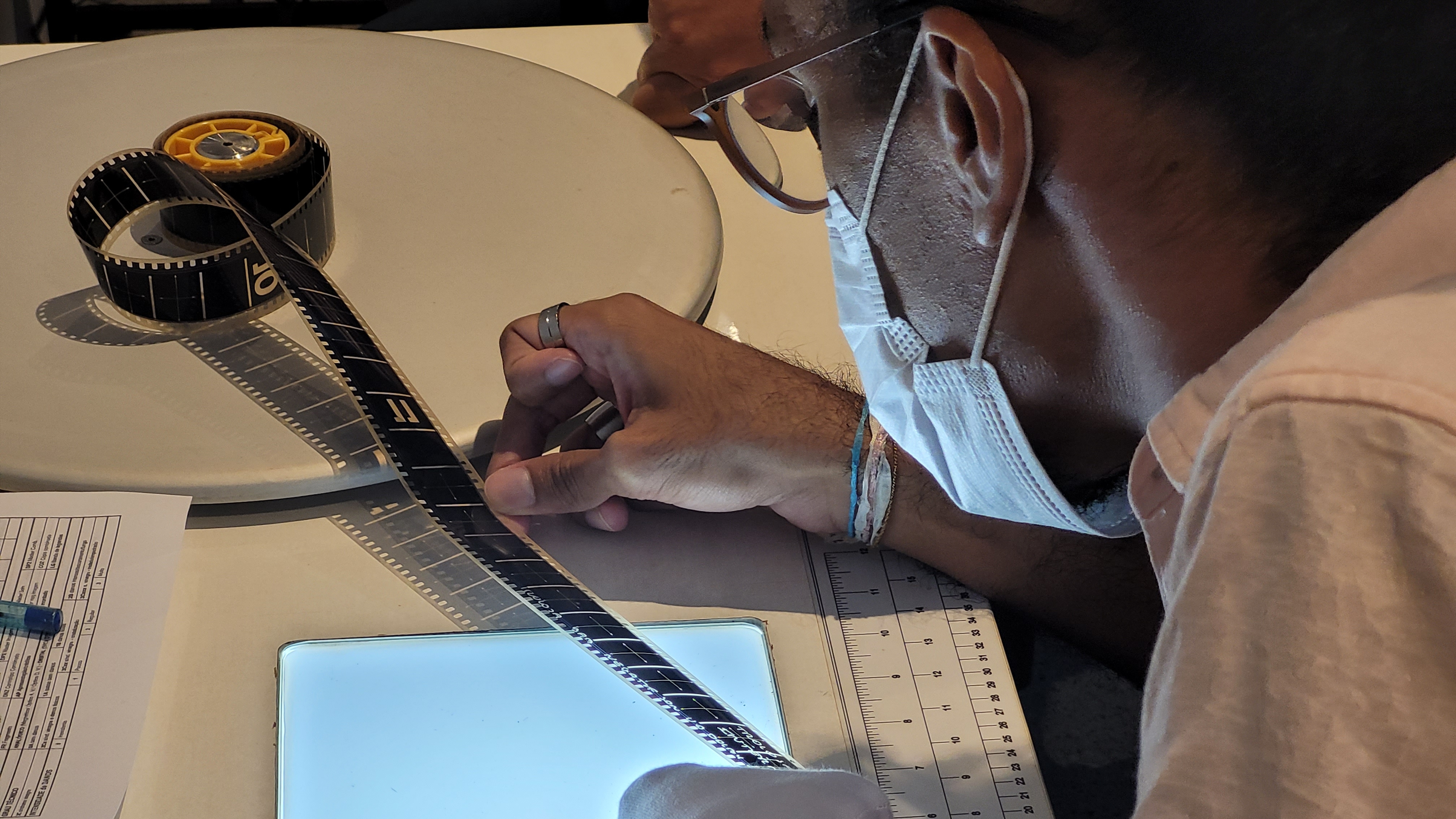 Um homem de perfil, usando óculos de grau e máscara cirúrgica branca, está inclinado sobre uma mesa de trabalho. Ele veste uma camisa rosa claro e tem no pulso um mix de pulseiras. Sua mão direita, com um anel prateado no dedo médio, segura e estica uma fita de filme de cinema preta sobre uma pequena mesa de luz quadrada e brilhante. O filme sai de um carretel de plástico amarelo que está sobre um grande disco branco mais acima na mesa. A mão esquerda dele está parcialmente coberta por uma luva branca e segura a ponta inferior do filme sobre a luz. Próximo à mesa de luz, à direita, há uma régua branca. À esquerda, sobre a mesa branca, vê-se um canto de uma folha de papel impressa com uma tabela e um objeto azul não identificado. A iluminação é focada na área de trabalho.